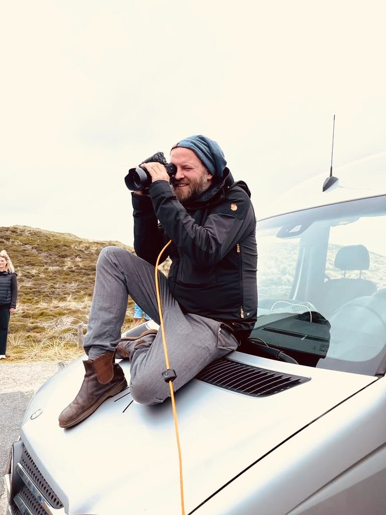Daniel Reinhold sitzt mit einer Kamera auf der Motorhaube eines Autos, dahinter Dünen